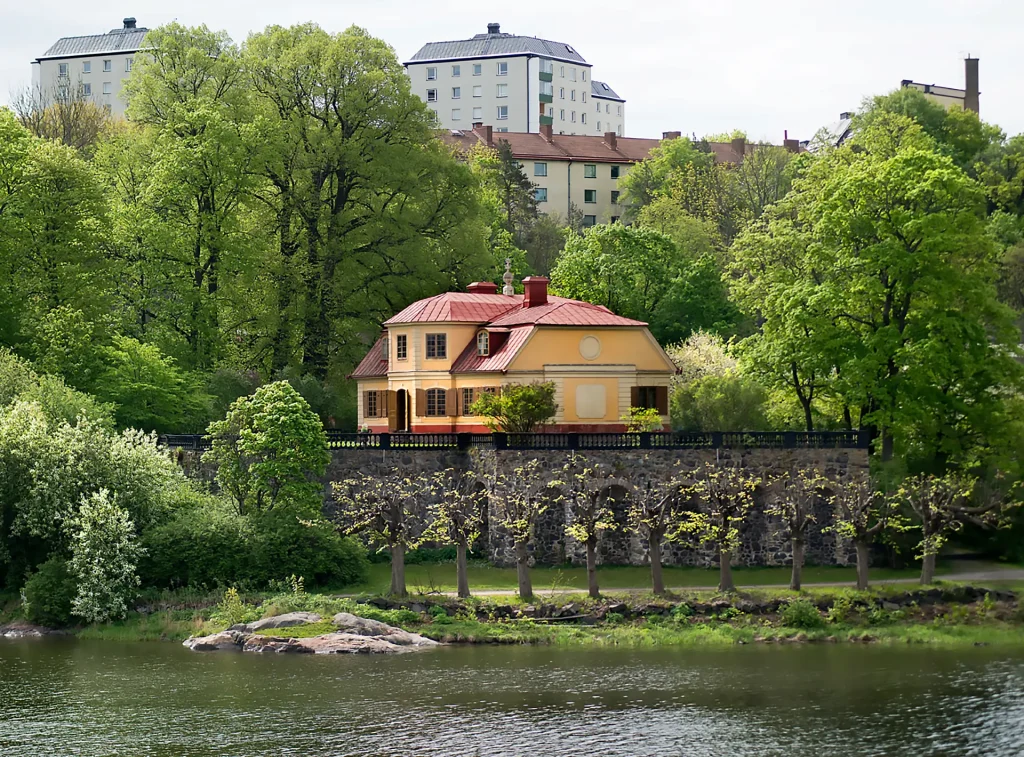Gul liten 1700-talsherrgård vid vatten, i bakgrunden syns moderna höghus.